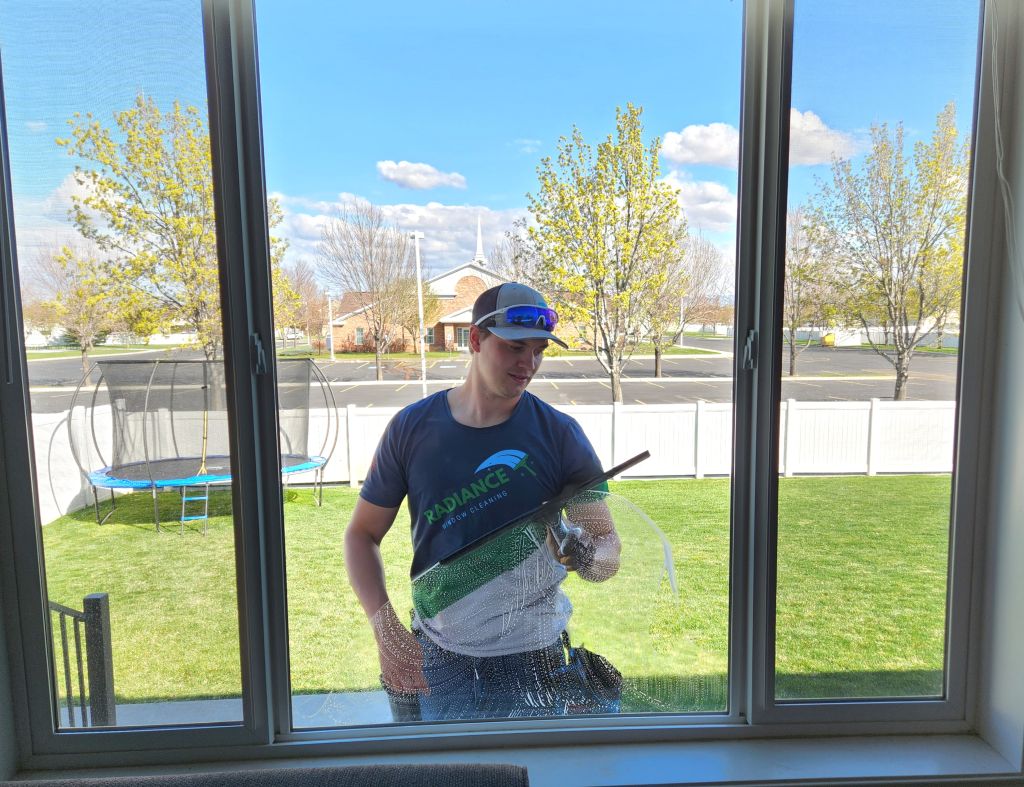 Man cleaning a large window from outside, squeegee in hand, wearing a cap and sunglasses; backyard with a trampoline and trees outside.