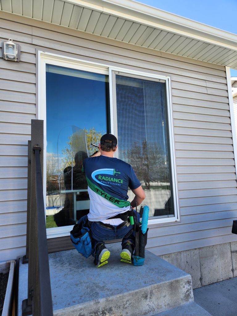 Window cleaner kneeling on exterior steps, scrubbing a large glass door with a squeegee; wearing a Radiance Window Cleaning shirt and tool belt.